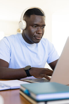 African American man sitting at desk wearing headphones and blue t-shirt focusing on laptop