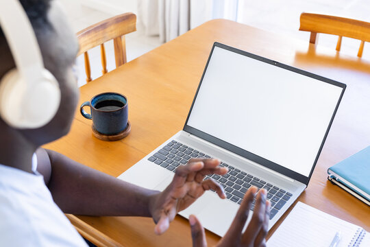 Person wearing white headphones gesturing at blank laptop screen on wooden table, copy space