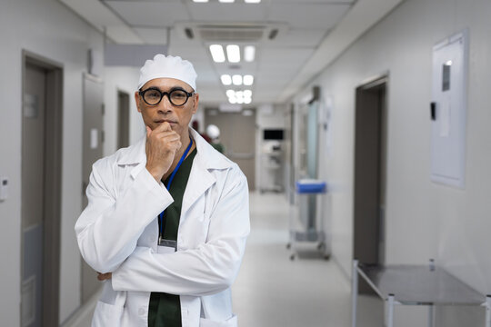 African American man in lab coat and scrubs standing with hand on chin in hospital corridor