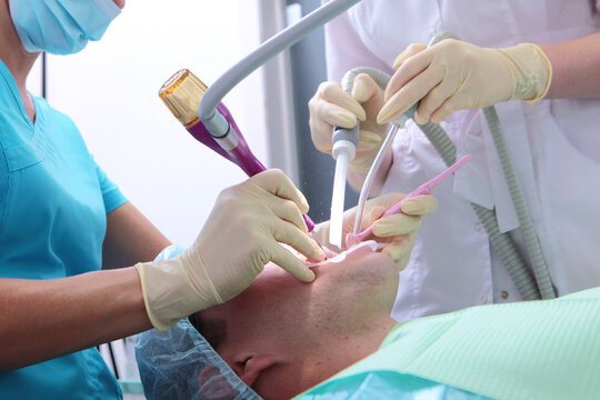 Dentist and assistant perform professional teeth cleaning for male patient. Air-polishing teeth whitening. Use dental instrument, suction, mirror, retractor. Clinical office, gloves, masks, bright lig