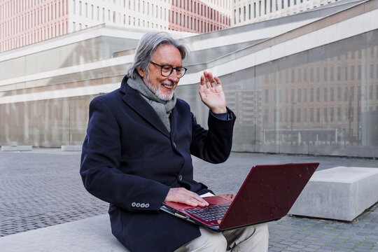 hispanic senior businessman using laptop sitting on bench outdoors in city, mature or elderly man working with computer in cityscape 