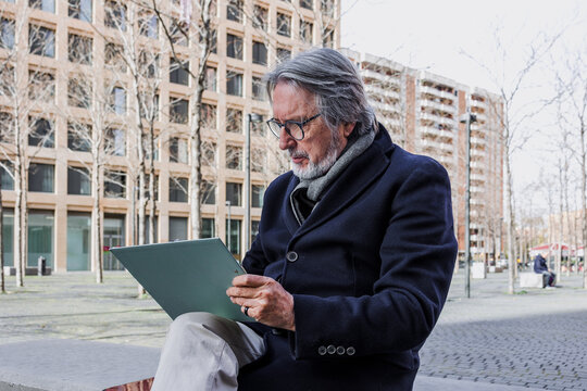 hispanic senior businessman using laptop sitting on bench outdoors in city, mature or elderly man working with computer in cityscape 