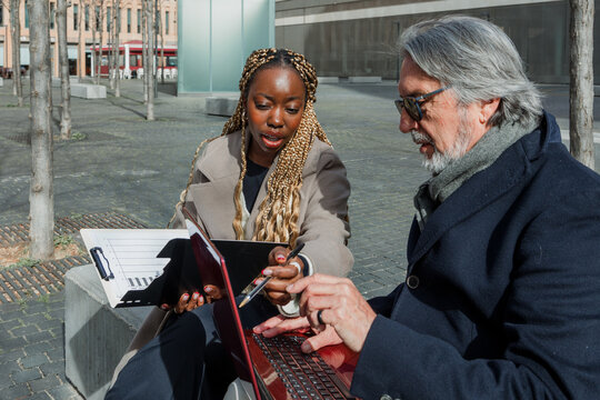 Young Adult black woman and Hispanic Business senior man shaking hands and using laptop Outdoors in a winter morning in cityscape. Manager and employee