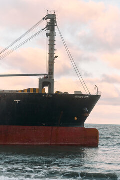 Large cargo ship anchored near shore at sunset with rusted red hull and black upper plate, deck cranes and rigging visible against pastel sky, calm sea and low waves in foreground suggesting coastal,.