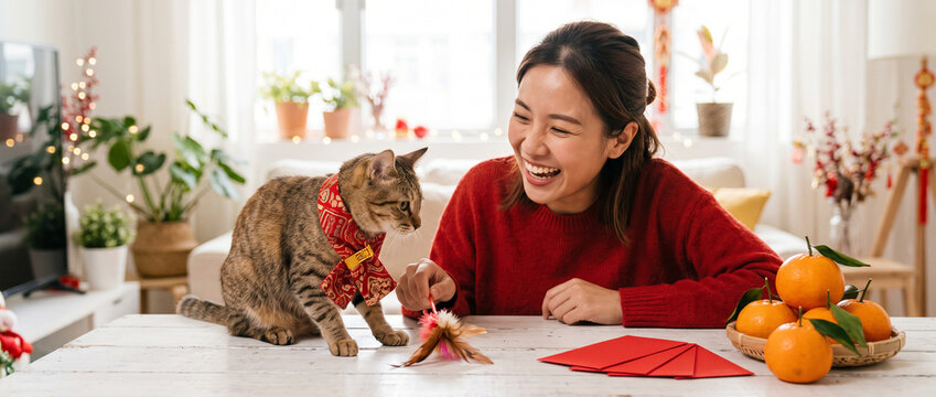 Asian woman in traditional red Chinese clothing playing with a cat beside red envelopes and orange decorations, Lunar New Year celebration and festive home