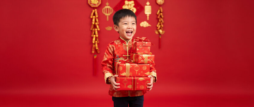 Happy Chinese boy in red traditional clothing carrying wrapped presents, Lunar New Year greeting concept, family celebration and good fortune