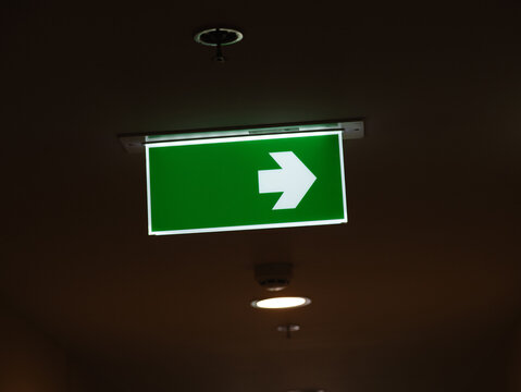 Illuminated Green Emergency Exit Direction Sign with White Right Arrow Hanging in Dark Indoor Corridor, Safety and Evacuation Concept