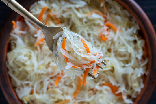 Sauerkraut with carrots on fork over ceramic bowl top view. Closeup shot of homemade fermented sauerkraut with shredded carrots on vintage fork held over traditional clay bowl
