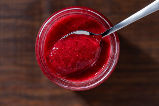 Spoonful of red berry jam in glass jar, closeup. Metal spoon scooping fresh red cranberry jam, puree or sauce from glass jar on dark wooden background, macro top view