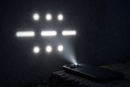 Smartphone flashlight beam projecting SOS in Morse code dots and dashes onto a plain dark wall at night in crisp light, a cry for help carried on a glowing screen in a quiet moment