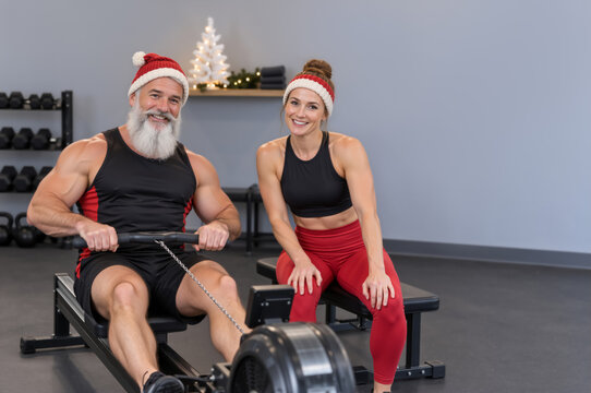 Fit Santa Claus using rowing machine with female workout partner in gym. Cheerful senior man and young woman exercising in Christmas hats