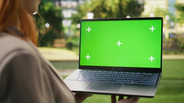 Businesswoman holds a green screen laptop mockup for website or advertising content in an urban park, presenting an isolated editable display suitable for replacing the screen digitally.