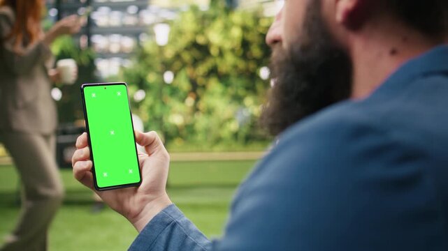 Man presents a phone with a vertical green screen mockup in an urban park, holding the isolated chroma key display for editable website, advertising or mobile app content replacement.