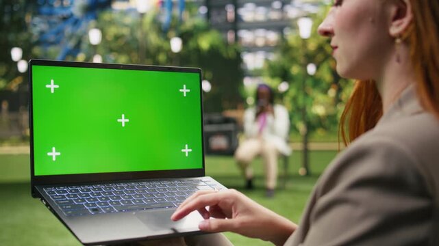 The laptop with an isolated green screen is held against a blurred city park, offering a blank editable display for digital screen replacement.