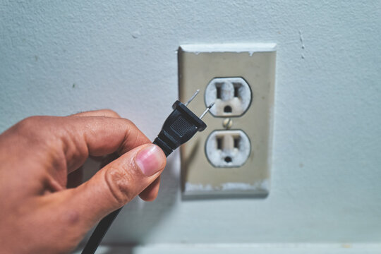 Hand Holding a Black Plug Near a Wall Electrical Outlet