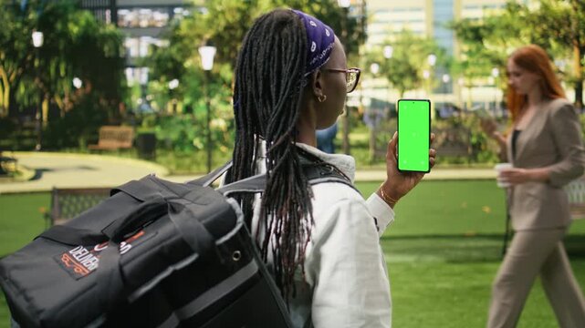 Food delivery courier presents a vertical chroma key smartphone near a bicycle in an urban park, providing a blank replaceable display for website, advertising or mobile app content.