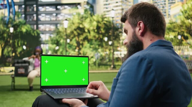 Businessman holds a green screen laptop mockup against a blurred city park, showing an isolated chroma key display ready for editable digital content.