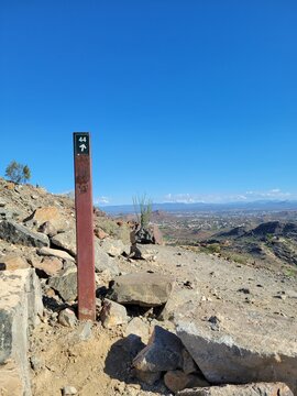 A weathered brown trail marker stands on a rocky desert peak offering a sweeping panoramic view of a valley city below under clear skies, North Mountain park, Phoenix, Arizona.