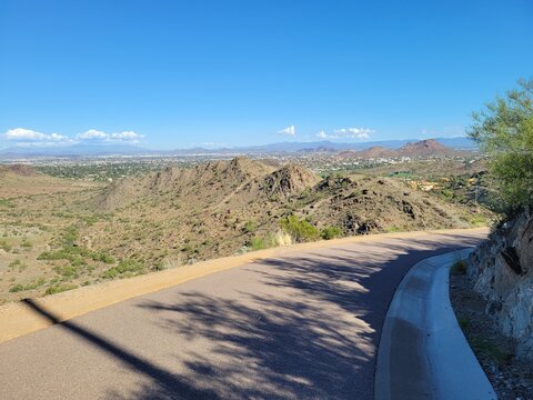 A smooth asphalt path curves down a steep rocky hillside, offering sweeping scenic vistas of arid mountains and a distant desert community under blue skies, North Mountain park, Phoenix, Arizona,
