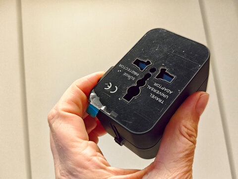 Hand holds a black universal travel adapter showing its multi-standard socket face labeled Travel Universal Adaptor and Surge Protector.