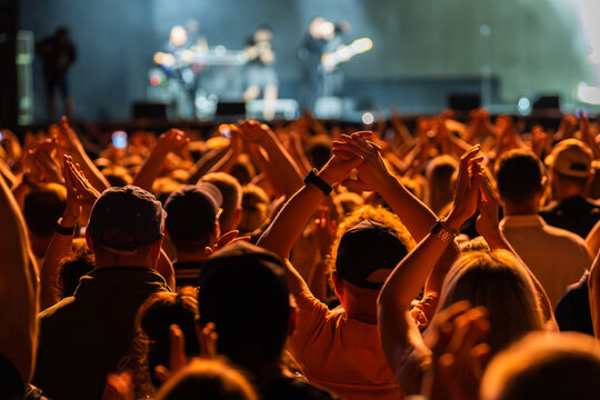Crowd of enthusiastic people enjoying a live music concert, raising their hands and cheering in front of a brightly illuminated stage at night.