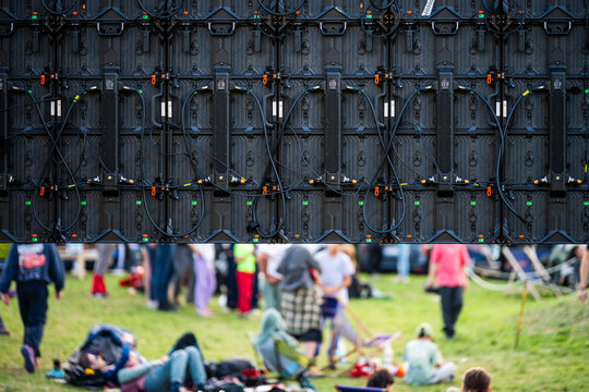 Backside view of a large modular LED screen wall with intricate wiring and cables at an outdoor festival with crowd resting on grass.