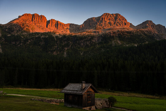 Majestic mountain peaks glowing in warm golden sunset light above dark pine forest in Dolomites, Italy.