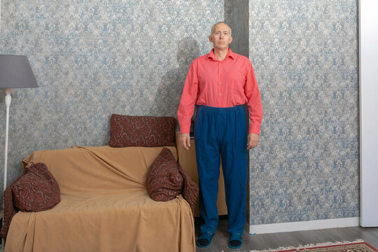 Man standing beside a covered sofa in a patterned living room. Interior portrait of an adult at home. Domestic life and everyday routine. Home furnishings and interior design