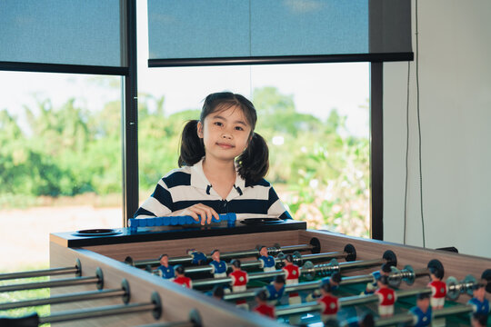 Young girl with ponytail playing foosball indoors near large window with greenery view in casual striped shirt