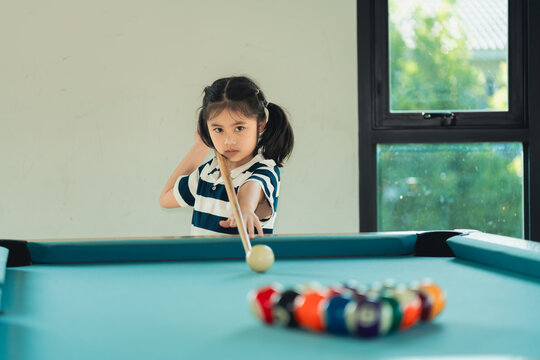Young girl concentrating on playing billiards aiming to strike cue ball on pool table in home setting