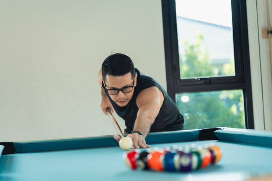Focused young man aiming with cue stick to strike the cue ball on a billiard table in bright modern room