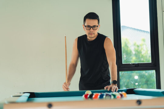 Focused young man wearing glasses and black sleeveless shirt playing pool indoors with window view and natural light