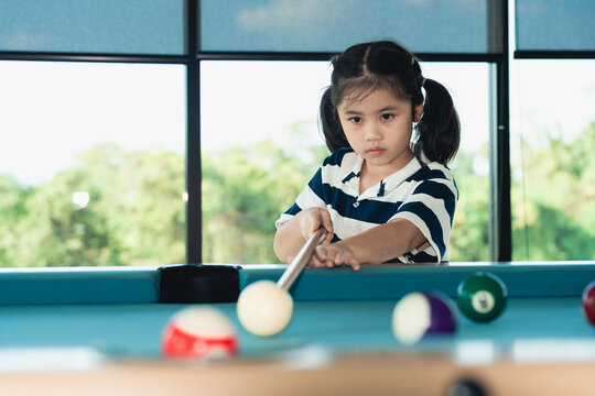 Young girl with serious expression aiming cue stick to strike white ball on billiard table in bright room