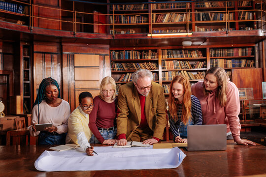 Professor guides university students studying architectural blueprints in a historic library