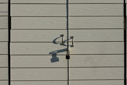 Light grey wooden door with two metal handles and black padlock. Strong shadows cast on surface. Locked gate. Minimalist closeup
