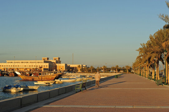 Person walking along a palm lined seaside promenade next to moored boats, Capital Governorate, Kuwait City, Kuwait