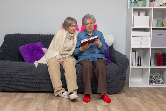 Two elderly women reading a book together on a sofa at home. Grandmothers sharing a quiet reading moment. Senior companionship and family connection. Leisure, mental health, dementia, Alzheimers