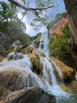 Vertical scenic view of the seventh tier of Erawan Waterfall in Kanchanaburi, Thailand. Cascading water descends a rocky cliff and flows over large natural rock formations surrounded by tropical veget