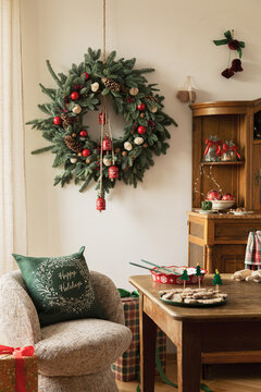 Cozy rustic Christmas interior with a large green fir wreath hanging on the wall, festive decor, wooden table, armchair with pillow, and holiday treats