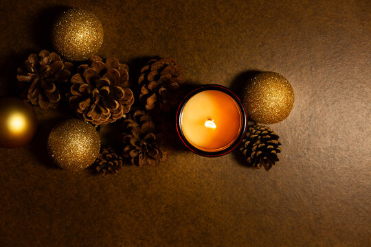 Bougie allum&eacute;e dans un pot ambr&eacute; entour&eacute;e de pommes de pin et de boules de No&euml;l dor&eacute;es et paillet&eacute;es, vue de dessus sur un fond brun