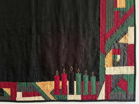 Kwanzaa quilted textile design featuring traditional fabric patchwork borders, embroidered kinara candles, black background field, and cultural heritage copy space for annual celebrations