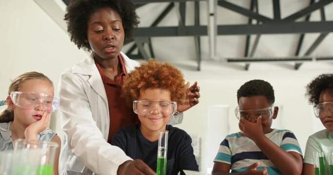 Diverse teacher with pupils guiding experiment at school lab, wearing lab coat, goggles, test tubes