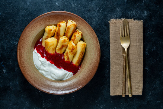 Crepes with sweet berry jam and sour cream on dark background top view, closeup. Rolled thin pancakes served with red berry sauce and sour cream in ceramic plate next to fork and knife on napkin