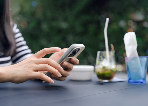 Woman using smartphone at outdoor cafe, hands texting and browsing online beside iced drink and garden background, casual lifestyle, mobile communication and relaxing spring summer leisure break.