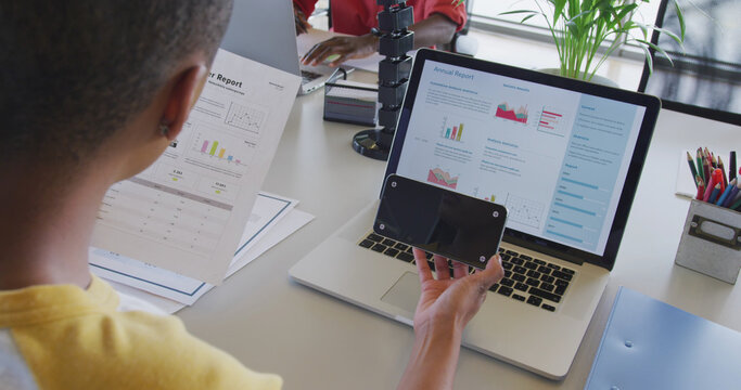African american businesswoman at desk, reading report, using laptop and smartphone with copy space