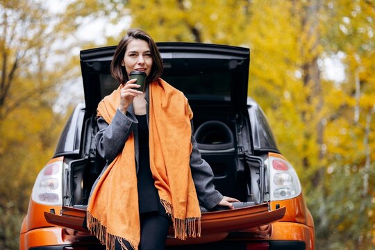 Woman sipping takeaway coffee beside an open car trunk, wrapped in an orange scarf amid golden autumn trees