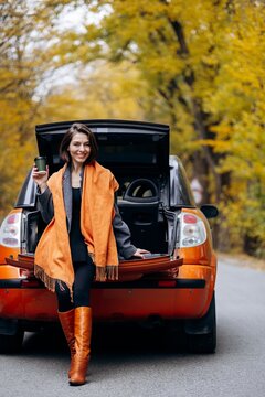 Woman traveler holding takeaway coffee while sitting on an open orange car trunk during an autumn road trip through fall foliage