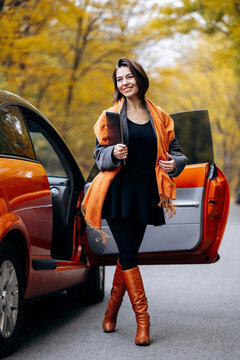 Professional woman standing beside an open orange car and holding a tablet, dressed for autumn among golden roadside trees