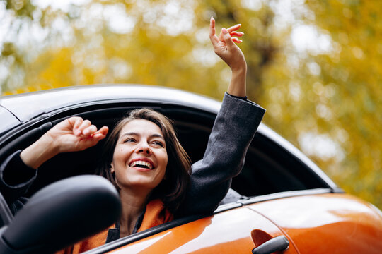 Smiling woman leaning from an orange car with arms raised, enjoying a carefree ride beneath golden autumn trees