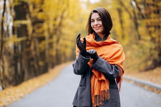 Woman in a gray coat and orange scarf adjusting her gloves on a quiet road amid golden autumn foliage in a wooded park